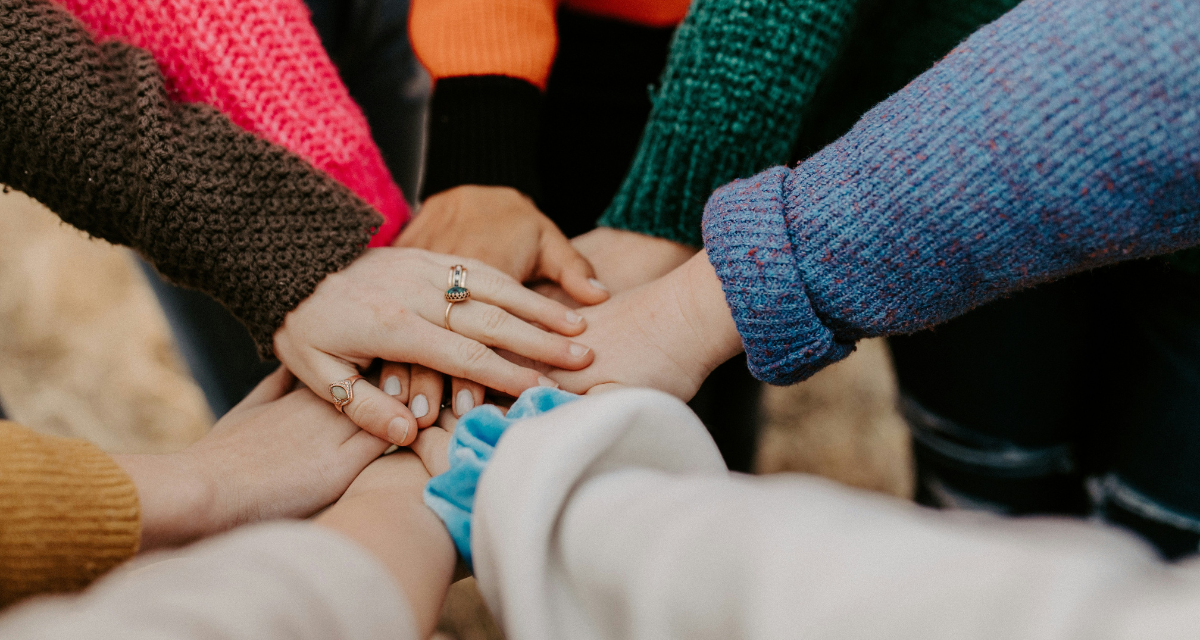 Gruppe stapelt Hände in bunte Pullover. Foto: Hannah Busing | Unsplash